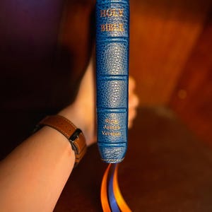 May include: A blue leather-bound Holy Bible with gold lettering. The spine reads "Holy Bible" and "King James Version." The book has three colored ribbon bookmarks. The image is taken from a low angle.