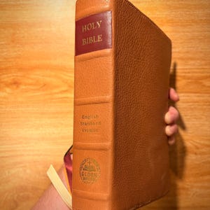 Puede incluir: Una Biblia encuadernada en cuero marrón, versión estándar en inglés, con letras doradas y lomo rojo. El libro tiene una cubierta texturizada y una cinta de marcador. Las palabras "Glory Bound" están impresas en la parte inferior del lomo.