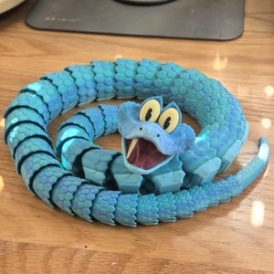 May include: A coiled, articulated toy snake in shades of blue, with a cartoonish face and open mouth. The snake's body is segmented, and the head features large, yellow eyes and a red mouth. The toy is on a wooden surface.