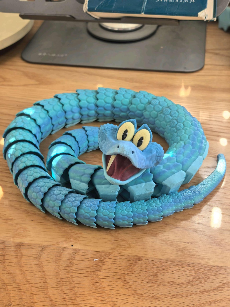 May include: A coiled, articulated toy snake in shades of blue, with a cartoonish face and open mouth. The snake's body is segmented, and the head features large, yellow eyes and a red mouth. The toy is on a wooden surface.
