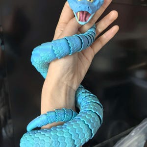May include: A blue articulated snake toy with a cartoonish head featuring yellow eyes and an open mouth. The body of the snake is a lighter shade of blue with a textured, scale-like pattern. The toy is flexible and appears to be made of plastic.