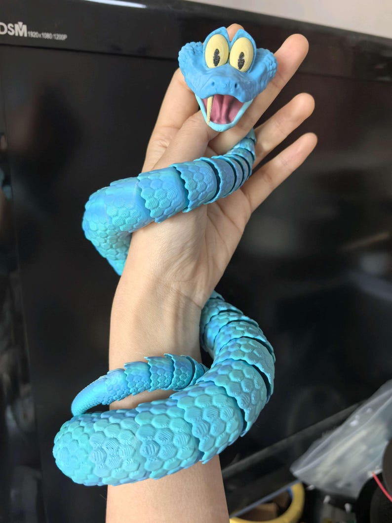 May include: A blue articulated snake toy with a cartoonish head featuring yellow eyes and an open mouth. The body of the snake is a lighter shade of blue with a textured, scale-like pattern. The toy is flexible and appears to be made of plastic.