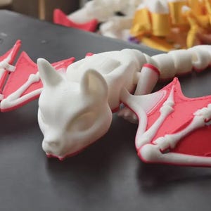 May include: A white 3D-printed cat-like creature with red and white wings. The model has a skeletal design, with visible bone structures in the wings and body. The creature is lying on a dark surface.