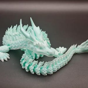 Op de afbeelding: Een lichtblauwe drakenfiguur met ingewikkelde details. De draak staat in een kronkelende positie, met zijn hoofd omhoog en staart gekruld. De figuur is gemaakt van een glad materiaal en heeft een getextureerd oppervlak.