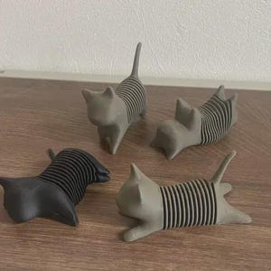 May include: Four cat-shaped figurines in varying poses. Three are gray, and one is black. Each cat has a striped design on its body. The figurines are displayed on a wooden surface.