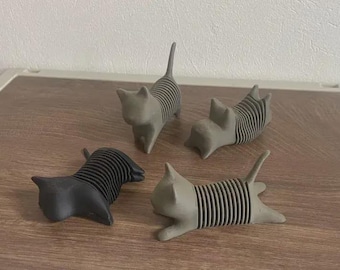 Puede incluir: Cuatro figuras con forma de gato en varias poses. Tres son grises y una es negra. Cada gato tiene un diseño a rayas en su cuerpo. Las figuras se exhiben sobre una superficie de madera.