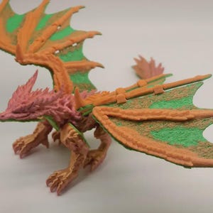 Puede incluir: Figura de dragón tridimensional detallada. El dragón presenta una cabeza y un cuerpo rosados, con alas naranjas y verdes. Las alas están extendidas y el dragón posa como si estuviera a punto de emprender el vuelo. La figura está hecha de un material texturizado.