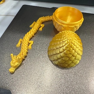 Pode incluir: Um dragão amarelo dourado impresso em 3D com detalhes intrincados, ao lado de um recipiente em forma de ovo correspondente e um recipiente redondo. O dragão está posado com o corpo curvado. O recipiente em forma de ovo tem uma superfície texturizada.