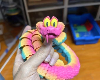 May include: A colorful, articulated snake toy in shades of pink, yellow, blue, and green. The snake has a cartoonish face with large yellow eyes and a pink tongue. The toy is held in a hand and appears to be made of plastic.