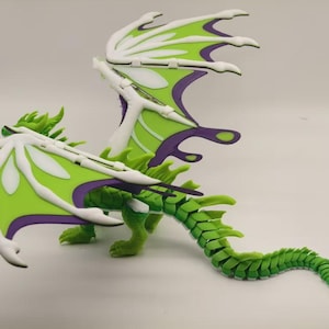 Op de afbeelding: Een groene en witte drakenfiguur met beweegbare vleugels en staart. De vleugels hebben een limoengroene basis met witte en paarse accenten. Het lichaam van de draak is groen en de staart is gesegmenteerd.