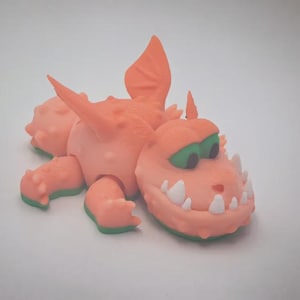 May include: A peach-colored dragon figurine with a textured, bumpy surface, green eyes, and white teeth. The creature features small wings on its back, a spiked tail, and four legs. The underside of the dragon is a solid green color.