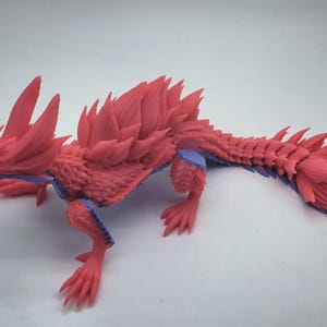 Op de afbeelding: Een 3D-geprinte, beweegbare drakenfiguur met een felrood lichaam en scherpe, gelaagde schubben langs de rug en staart. Het wezen heeft een vossenachtige kop, vier ledematen met klauwen en contrasterende blauwe gewrichten bij de schouders en heupen.