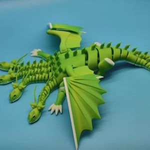 Op de afbeelding: Een groene, meerkoppige drakenfiguur met witte accenten op een blauwe achtergrond. De draak heeft grote vleugels en een gesegmenteerd lichaam. De vleugels van de draak zijn een lichtere tint groen.