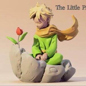 May include: A figurine of The Little Prince, a character from the classic children's book. The figure is depicted in a green outfit with a yellow scarf and hair, seated on a stone-like structure, looking at a red rose. The text "The Little Prince" is above the figure.