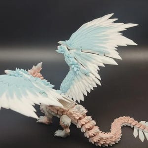 May include: A detailed figurine of a mythical creature with large, feathered wings. The wings are primarily white with light blue accents. The body is a mix of light pink and beige, with a textured, scaled appearance. The creature has a long, segmented tail.