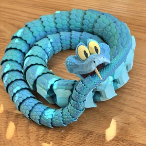 May include: A blue articulated snake toy with a cartoonish design. The snake is coiled, with a detailed scale pattern and a smiling face with large yellow eyes. The toy is made of plastic and appears to be 3D-printed.