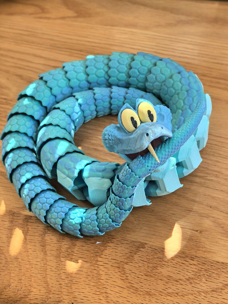 May include: A blue articulated snake toy with a cartoonish design. The snake is coiled, with a detailed scale pattern and a smiling face with large yellow eyes. The toy is made of plastic and appears to be 3D-printed.
