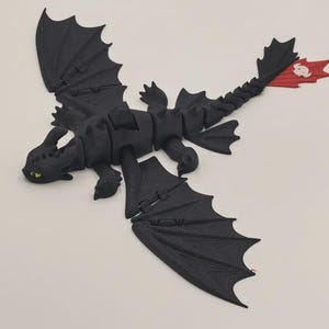 May include: A black articulated dragon figurine with large wings, a long segmented tail, and a red tail fin featuring a white skull-like emblem. The dragon has green eyes and a textured surface, depicted in a flying pose with limbs extended.
