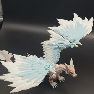 May include: A detailed figurine of a dragon with large, feathered wings. The dragon features a color palette of white, light blue, and pale pink. The wings are spread, and the dragon is posed on a dark surface.