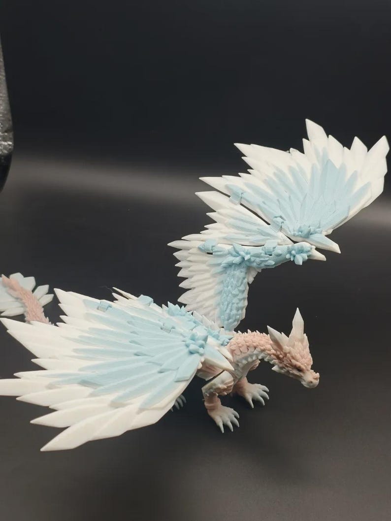 May include: A detailed figurine of a dragon with large, feathered wings. The dragon features a color palette of white, light blue, and pale pink. The wings are spread, and the dragon is posed on a dark surface.