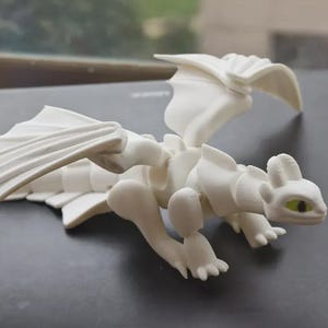Könnte beinhalten: Eine weiße Drachenfigur mit beweglichen Gliedmaßen, Flügeln und einem detaillierten Schwanz. Der Drache hat ein stilisiertes Design mit sichtbaren Gelenken und einem kleinen Kopf mit gelben Augen. Die Figur besteht aus einem glatten, matten Material.