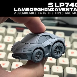 Op de afbeelding: Een grijze, in elkaar te zetten speelgoedauto, een Lamborghini Aventador, met beweegbare banden. De auto wordt in een hand gehouden, met een toetsenbord op de achtergrond. De tekst "SLP740-4 LAMBORGHINI AVENTADOR" staat bovenaan.