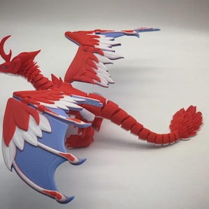 Op de afbeelding: Een rood, wit en blauw drakenbeeldje met scharnierende gewrichten. De draak heeft grote vleugels met blauwe onderkanten en witte en rode accenten. De staart is veervormig rood en de draak heeft een gebogen hoorn.