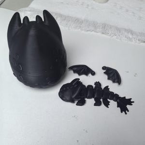 Op de afbeelding: Een zwarte 3D-geprinte drakenfiguur met afneembare vleugels en een gesegmenteerd lichaam. De drakenkop heeft prominente oren en gedetailleerde texturen. De vleugels zijn gespreid en het lichaam is gearticuleerd.