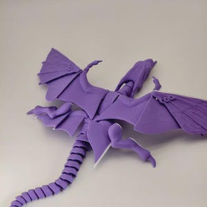 Könnte beinhalten: Eine violette, bewegliche Drachenfigur mit detaillierten Flügeln, Beinen und einem segmentierten Schwanz. Der Drache ist in einer Flugpose dargestellt, mit weit ausgebreiteten Flügeln. Die Figur besteht aus einem glatten, festen Material.