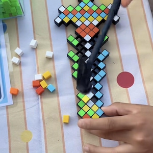Op de afbeelding: Een pixelachtig zwaardconstructiespeelgoed bestaande uit een zwart rasterframe gevuld met kleine, vierkante, veelkleurige tegels in tinten groen, oranje, geel, wit en lichtblauw. Een zwart magnetisch hulpmiddel wordt gebruikt om de tegels te plaatsen.