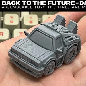 May include: A gray 3D-printed toy car, a DMC-12 from Back to the Future, held in a hand. The text "BACK TO THE FUTURE - DMC-12" and "ASSEMBLEABLE TOYS THE TIRES ARE MOVABLE!" is visible. The car has detailed features and movable tires.