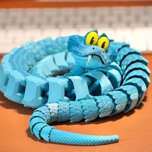 May include: A coiled, articulated snake figurine in shades of blue. The snake has a cartoonish design with large yellow eyes, a white fang, and a segmented body. The toy is resting on an orange surface, with a keyboard in the background.