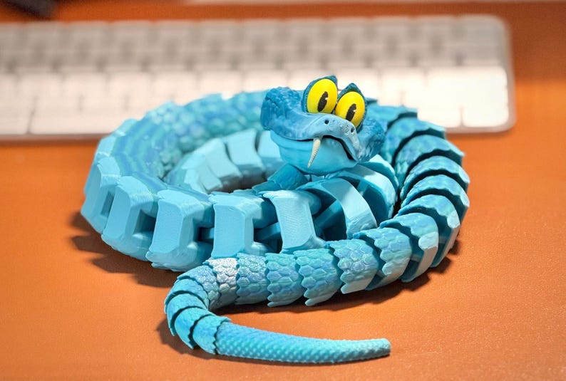 May include: A coiled, articulated snake figurine in shades of blue. The snake has a cartoonish design with large yellow eyes, a white fang, and a segmented body. The toy is resting on an orange surface, with a keyboard in the background.