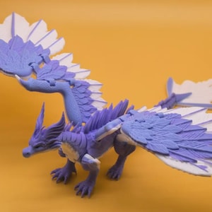 Op de afbeelding: Gedetailleerd drakenbeeldje, voornamelijk paars met witte vleugelaccenten. De vleugels zijn groot en waaiervormig. De draak staat op alle vier poten, met een gedetailleerde kop en staart. Gele achtergrond.