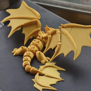 May include: A 3D-printed articulated dragon figurine in a light yellow color. The dragon has detailed wings, a segmented body, and a long tail. The figurine is posed on a dark surface, showcasing its intricate design and structure.