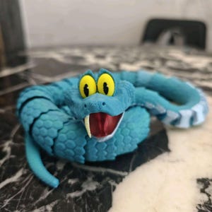 May include: A turquoise toy snake with cartoonish features. The snake has large yellow eyes, a red mouth with a white fang, and black stripes along its back. The toy is made of a textured material and is posed in a coiled position.