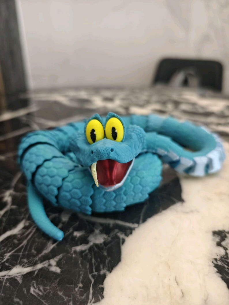 May include: A turquoise toy snake with cartoonish features. The snake has large yellow eyes, a red mouth with a white fang, and black stripes along its back. The toy is made of a textured material and is posed in a coiled position.