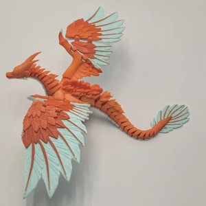 Op de afbeelding: Een gearticuleerd drakenfiguur met een gesegmenteerd lichaam met oranje schubben en lichtblauwe vleugeltips en staartvin. Het wezen staat in een vliegende positie met gespreide vleugels, met een gedetailleerd gelaagd verenontwerp op de vleugels.