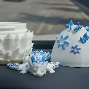May include: A collection of white 3D-printed fantasy figurines. The set includes a dragon, a domed structure with blue accents, and a decorative container with a crystal-like design.