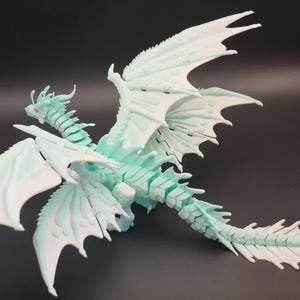 Puede incluir: Figura detallada de dragón en azul claro y blanco, con grandes alas de diseño intrincado. El dragón tiene una larga cola y un cuerpo detallado, con un aspecto texturizado. La figura se encuentra sobre un fondo oscuro.