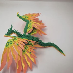 May include: A multi-jointed dragon figurine featuring a dark green segmented body and head. The dragon has large, expansive wings with layered, flame-shaped feathers in shades of yellow, light orange, and peach, accented with green circular patterns.