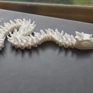 Op de afbeelding: Een witte, gelede drakenfiguur met een gedetailleerde, stekelige ruggengraat en een drakenkop. De figuur is gemaakt van een glad, stevig materiaal en staat op een donkere ondergrond. De drakenkop heeft zichtbare oogkassen en een gedetailleerde snuit.