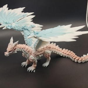 May include: A detailed figurine of a dragon with a light pink and white body, and large wings with blue and white accents. The dragon is posed with its wings spread, showcasing intricate scale and feather details. The figurine is on a dark surface.
