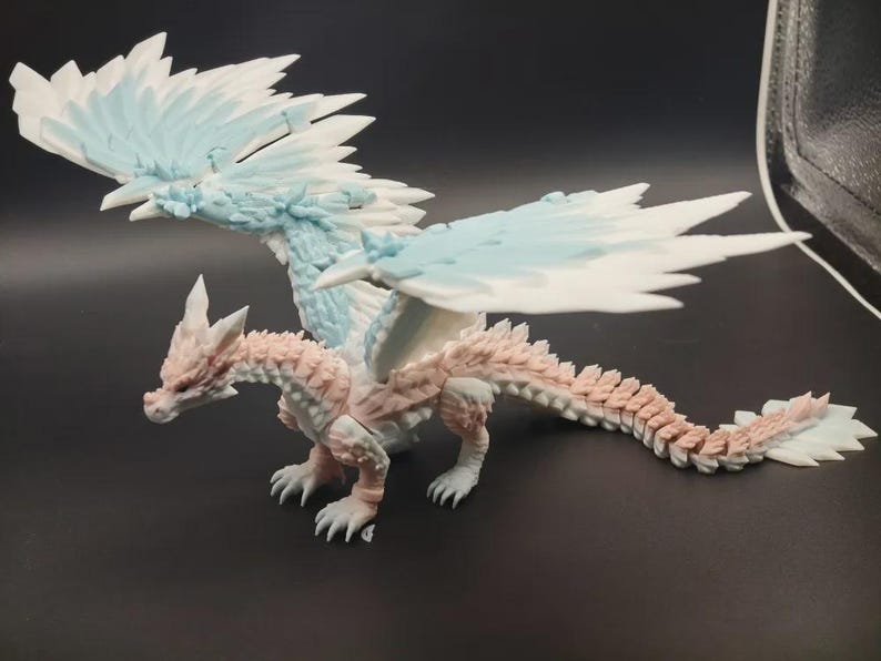 May include: A detailed figurine of a dragon with a light pink and white body, and large wings with blue and white accents. The dragon is posed with its wings spread, showcasing intricate scale and feather details. The figurine is on a dark surface.