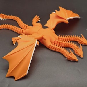 May include: An orange, three-headed dragon figurine with outstretched wings and multiple tails. The wings and tail tips have white accents. The dragon is posed on a dark gray surface.