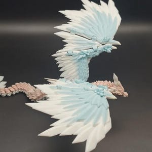 May include: A detailed, articulated dragon figurine with white, light blue, and pink accents. The dragon has large, layered wings and a long, segmented tail. The wings are spread, and the dragon appears to be in flight. The figurine is made of plastic.