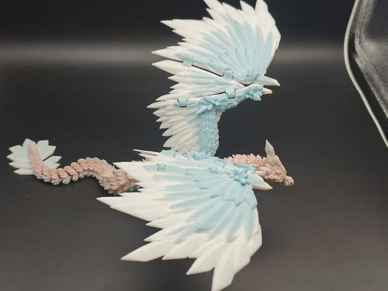 May include: A detailed, articulated dragon figurine with white, light blue, and pink accents. The dragon has large, layered wings and a long, segmented tail. The wings are spread, and the dragon appears to be in flight. The figurine is made of plastic.