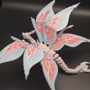 May include: A detailed, 3D-printed sculpture of a stylized insect-like creature. The wings are light blue with coral-colored accents and intricate patterns. The body and tail are a light gray color, with a segmented design. The sculpture is set against a dark background.