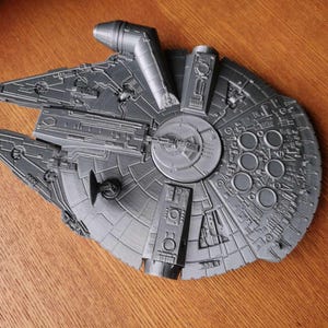Puede incluir: Un modelo gris detallado de la nave espacial Millennium Falcon. El modelo presenta intrincados paneles y detalles circulares, con un disco circular central. La nave está sobre una superficie de madera.