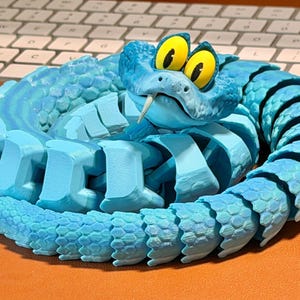 May include: A coiled, articulated blue snake toy with a detailed, textured design. The snake features large yellow eyes with black pupils and a forked tongue. The toy is made of plastic and sits on an orange surface.